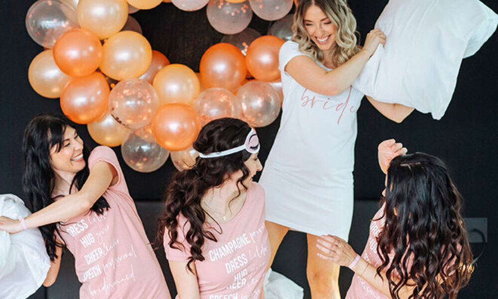 Pyjama-Party zum Junggesellinnenabschied: Die Braut in einem weißen Nachthemd und ihre Freundinnen in rosafarbenen Outfits haben Spaß bei einer Kissenschlacht, umgeben von einer dekorativen Ballonwand in Gold und Rosé.