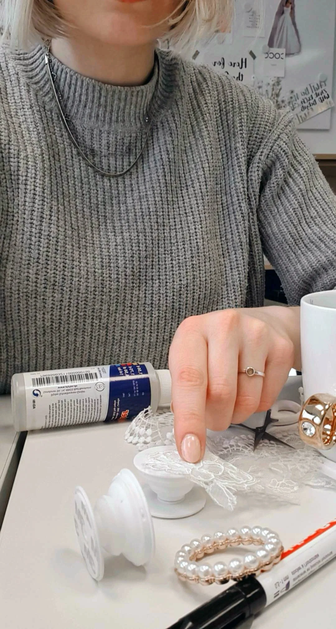 Eine Frau in einem Strickpullover arbeitet an einem Popsocket-DIY-Projekt und verwendet Spitze zur Verzierung. Weitere Bastelmaterialien wie Kleber und Perlen sind daneben arrangiert.