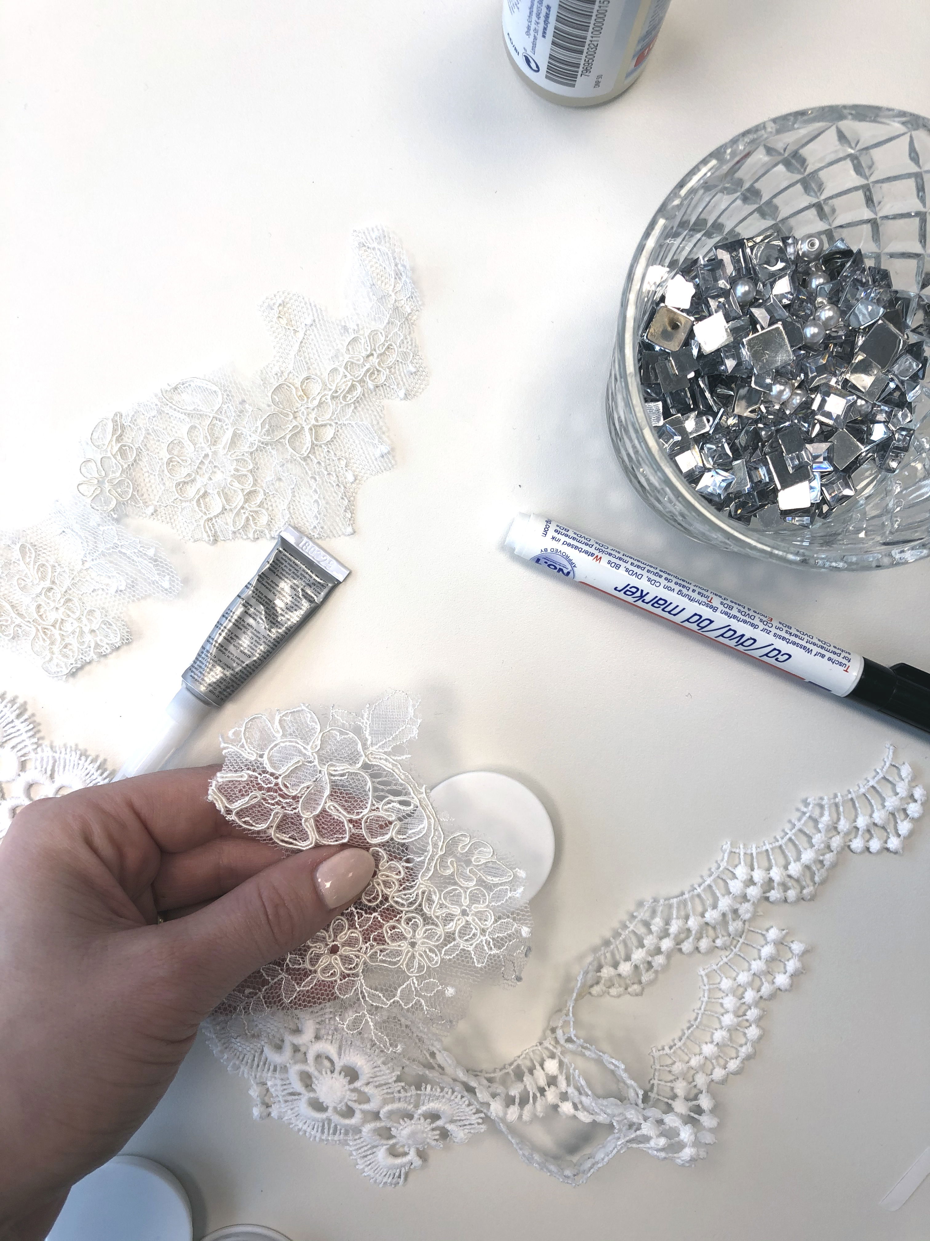 Eine Person greift nach einem Stück floral gemusterter Spitze für ein DIY-Projekt, umgeben von Kleber, glänzenden Steinchen und weiteren Bastelmaterialien auf weißem Hintergrund.