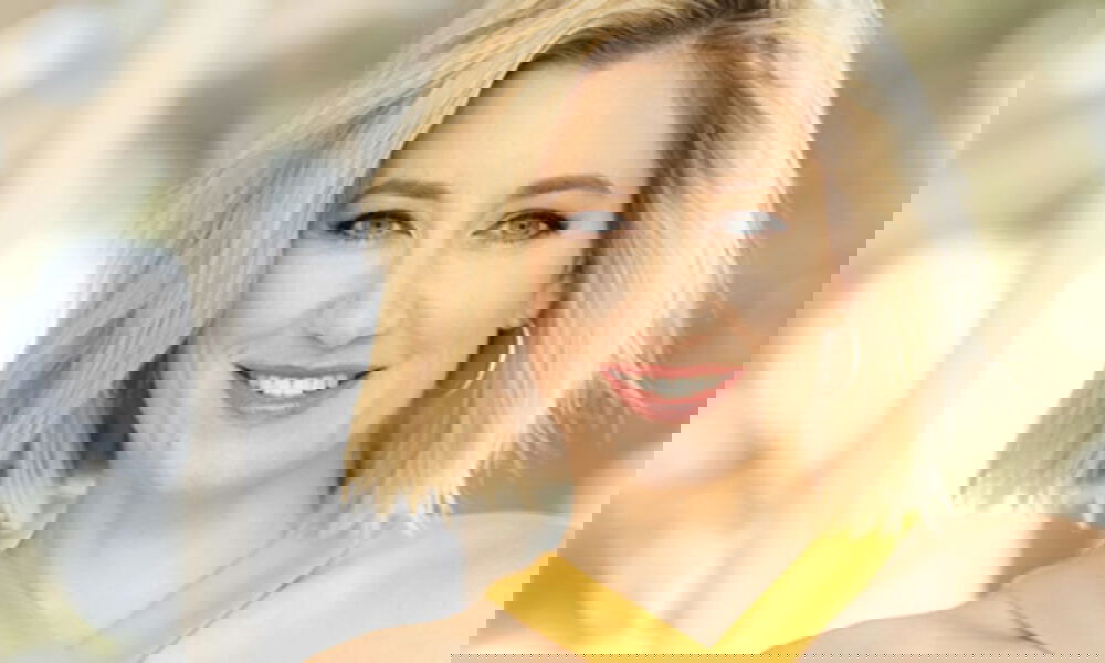 Blonde Frau mit kinnlangem Haar und einem freundlichen Lächeln blickt in die Kamera. Sie trägt ein Oberteil in warmem Gelb und dezenten Schmuck. Der Hintergrund ist weich und unscharf, was den Fokus auf ihr Gesicht lenkt.