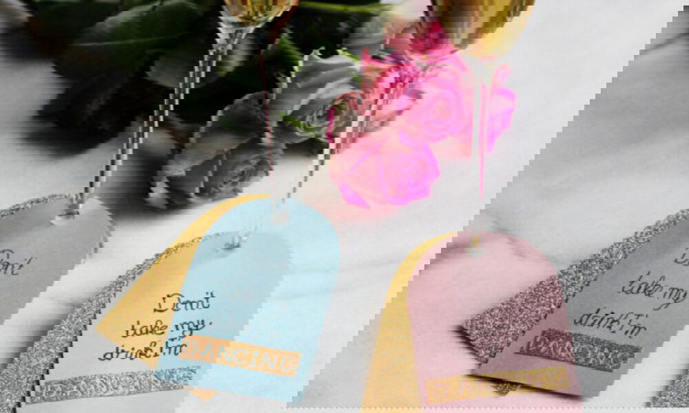 Zwei Sektgläser mit dekorativen Hangtags in Blau und Rosa, verziert mit goldenen Details und dem Text "Don’t take my drink, I’m dancing". Rosa Rosen sind im Hintergrund platziert.
