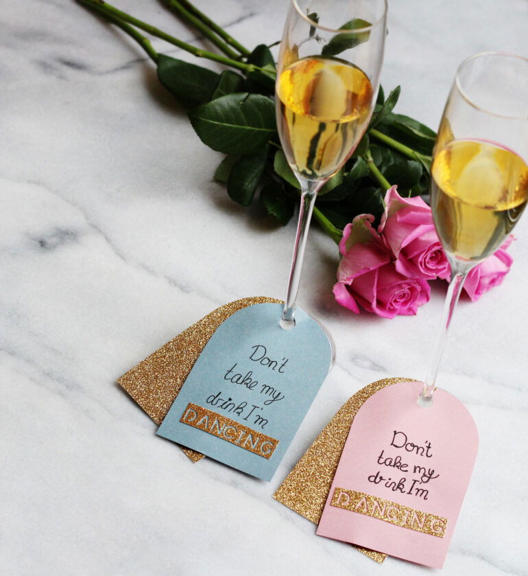 DIY-Hangtags an zwei Sektgläsern mit dem Text "Don’t take my drink, I’m dancing". Die Anhänger in Pastellfarben und mit Goldakzenten verleihen eine individuelle Note. Ein Rosenstrauß liegt im Hintergrund.