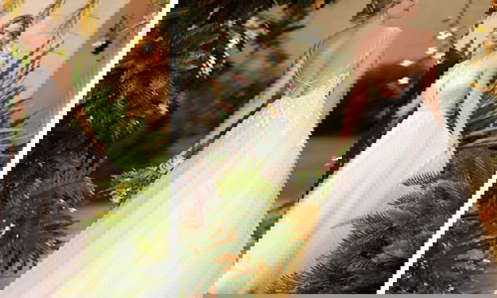 Links: Braut in einem ärmellosen Kleid mit Spitzendetails und weitem Rock, die Hand ihres Bräutigams haltend, in einem Garten voller exotischer Pflanzen. Rechts: Braut im Kleid mit offenem Rücken und filigranem Trägerdesign, inmitten tropischer Blätter bei sanfter Beleuchtung.