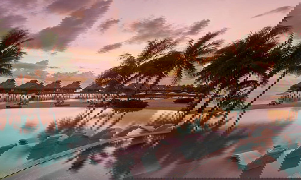 Infinity Pool mit Liegen im Wasser, eingerahmt von Palmen und Bungalows, in einer tropischen Landschaft bei Sonnenuntergang.