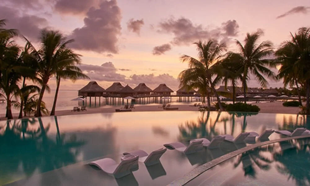 Infinity Pool mit Liegen im Wasser, eingerahmt von Palmen und Bungalows, in einer tropischen Landschaft bei Sonnenuntergang.