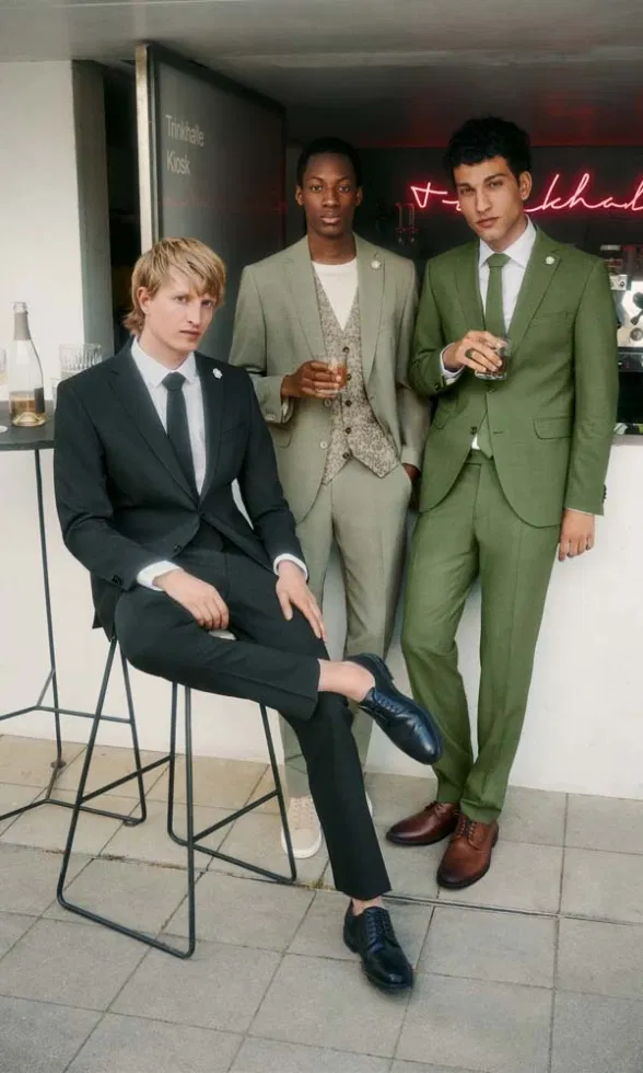 Drei Männer in modernen Anzügen in Grün- und Beigetönen stehen entspannt an einer Bar und halten Drinks in der Hand – ein urbanes Fashion-Shooting mit Hochzeitsbezug.