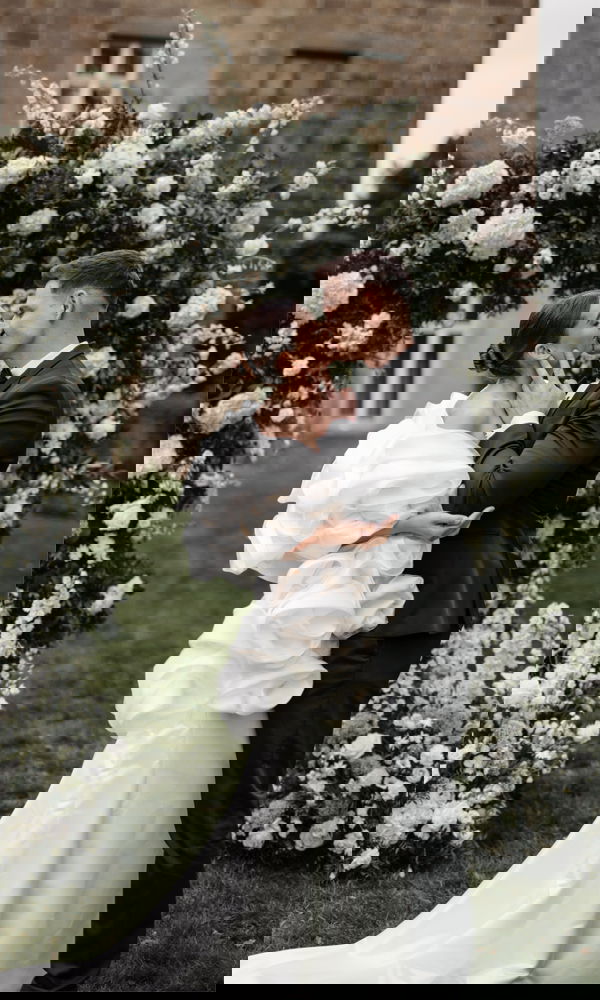 Hochzeitsfoto eines sich küssenden Brautpaares vor einem üppigen Blumenbogen aus weißen Blumen, die Braut trägt ein Kleid mit dramatischen Stoffdetails.