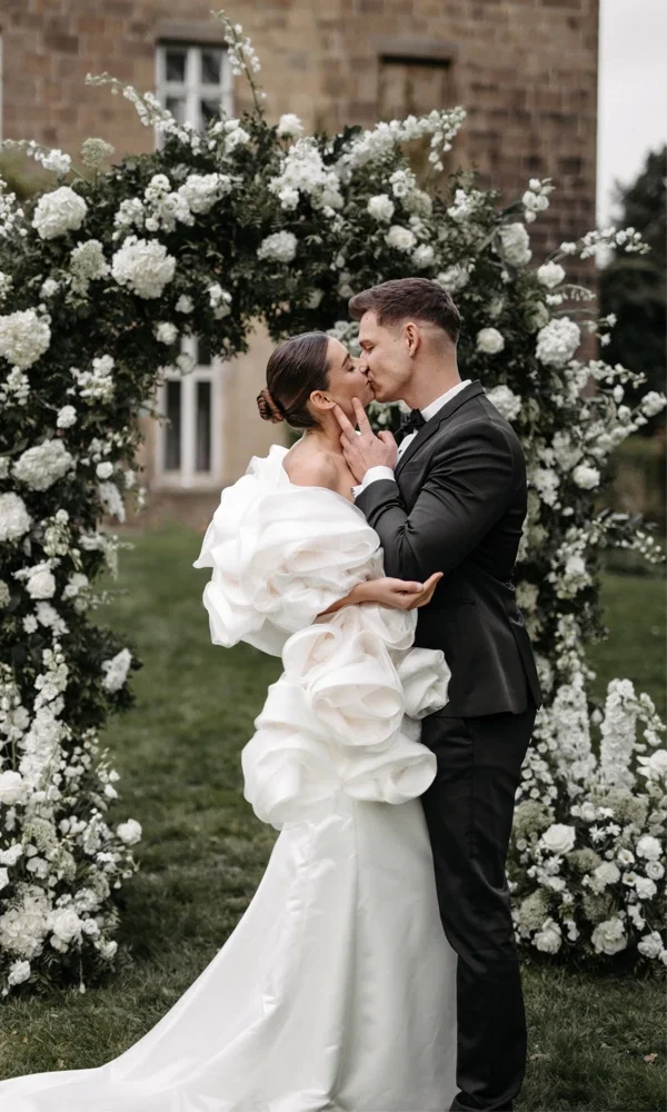 Hochzeitsfoto eines sich küssenden Brautpaares vor einem üppigen Blumenbogen aus weißen Blumen, die Braut trägt ein Kleid mit dramatischen Stoffdetails.
