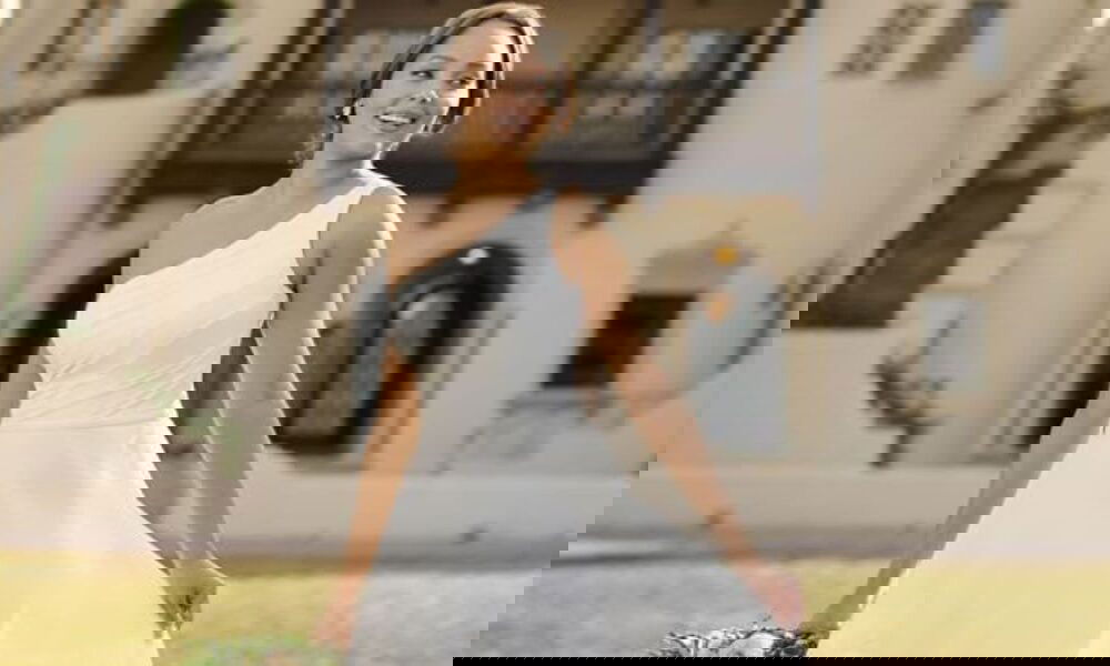 Junge Braut in einem schlichten, eleganten Kleid mit einem Träger, posiert fröhlich vor einem Herrenhaus. Der einseitige Schulterträger und der leichte Stoff unterstreichen den klassischen Look.