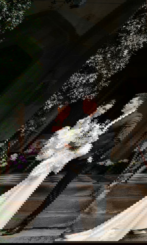 Braut mit großem, weißen Blumenstrauß und voluminösem Kleid steht neben Bräutigam im schwarzen Anzug auf den Stufen eines alten Gebäudes.