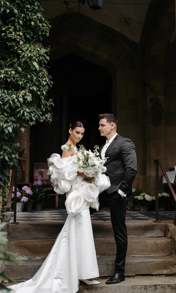 Braut mit großem, weißen Blumenstrauß und voluminösem Kleid steht neben Bräutigam im schwarzen Anzug auf den Stufen eines alten Gebäudes.