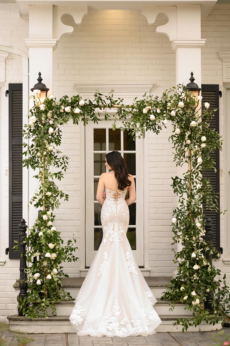 Eine Frau in einem trägerlosen, bestickten Brautkleid vor einer eleganten weißen Eingangstür, eingerahmt von grünem Blattwerk und weißen Blumen.