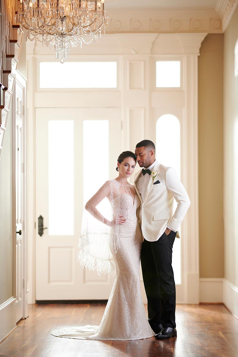 Das Brautpaar posiert in einem eleganten, hellen Raum, die Braut in funkelndem Kleid mit Fransen, der Bräutigam im weißen Smoking unter einem prächtigen Kronleuchter.