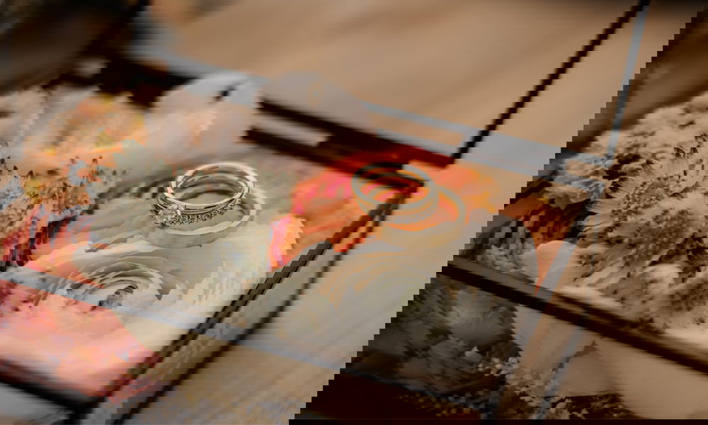 Dekorative Schmuckschatulle aus Glas, mit zarten Blumen in Rosa und Weiß gefüllt, auf denen die Hochzeitsringe kunstvoll platziert sind.