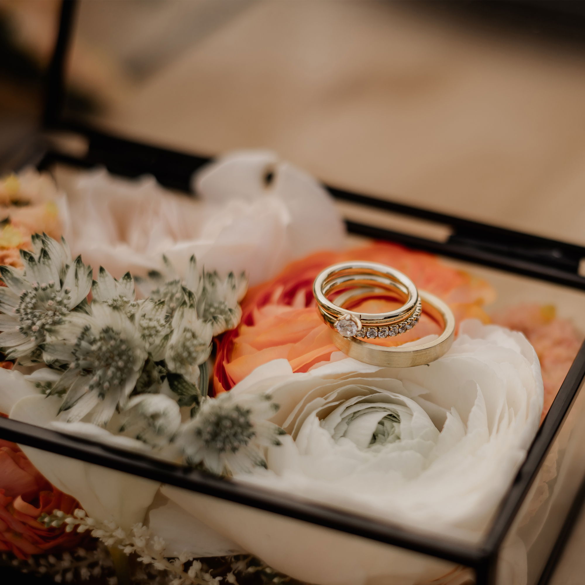 In einer transparenten Schmuckschatulle liegen die Hochzeitsringe auf einem Arrangement aus weißen und orangefarbenen Blüten, perfekt in Szene gesetzt.