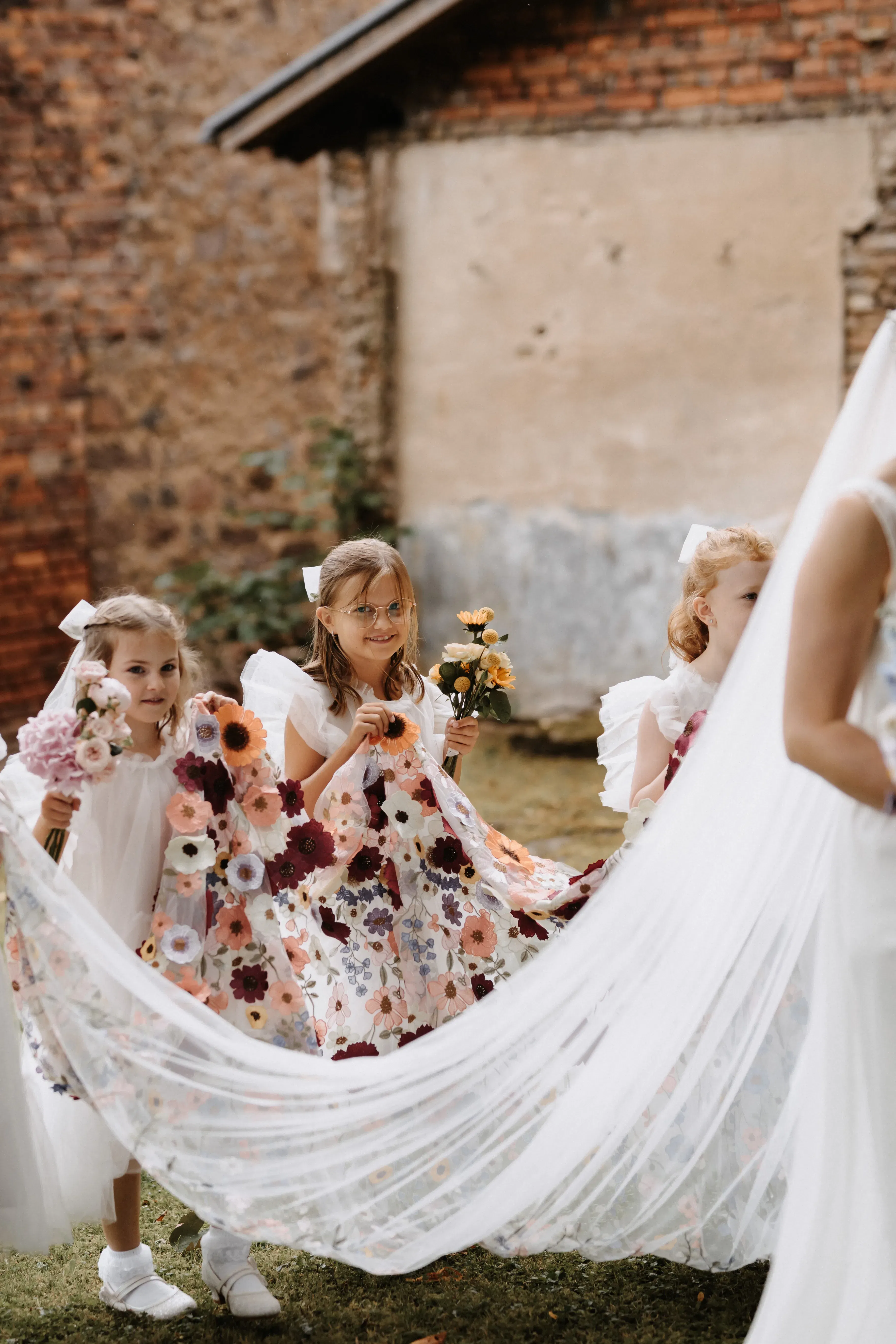 Drei Blumenmädchen in farbenfrohen, blütenverzierten Kleidern halten den langen Schleier der Braut während des Einzugs.