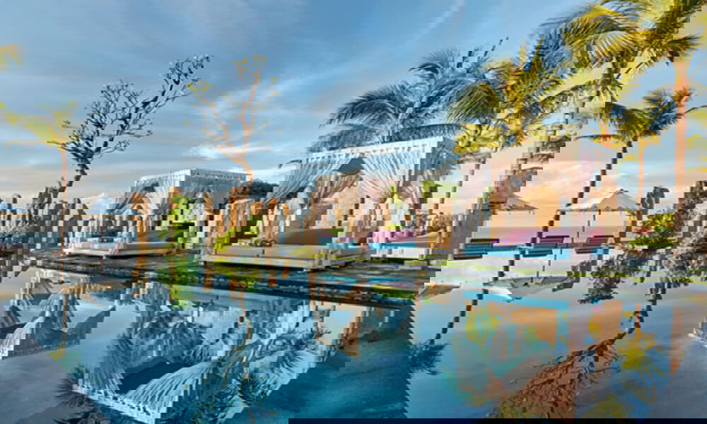 Eleganter Pool mit Spiegelung von Tagesbetten, umgeben von Palmen und Natursteinen unter klarem Himmel in einem Bali-Resort.