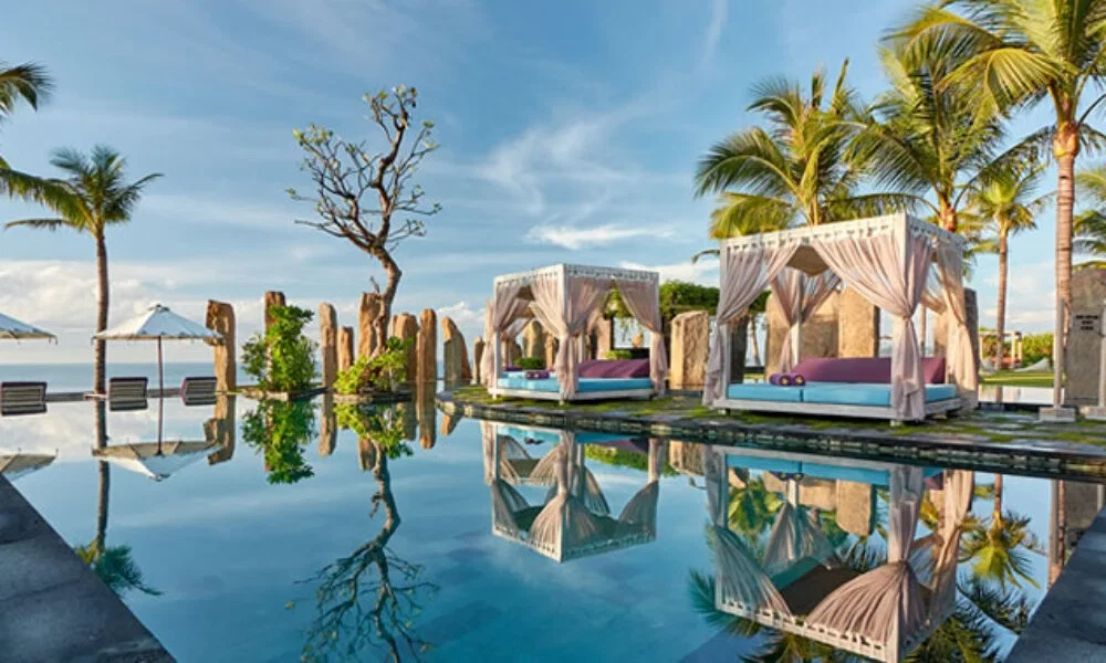 Eleganter Pool mit Spiegelung von Tagesbetten, umgeben von Palmen und Natursteinen unter klarem Himmel in einem Bali-Resort.