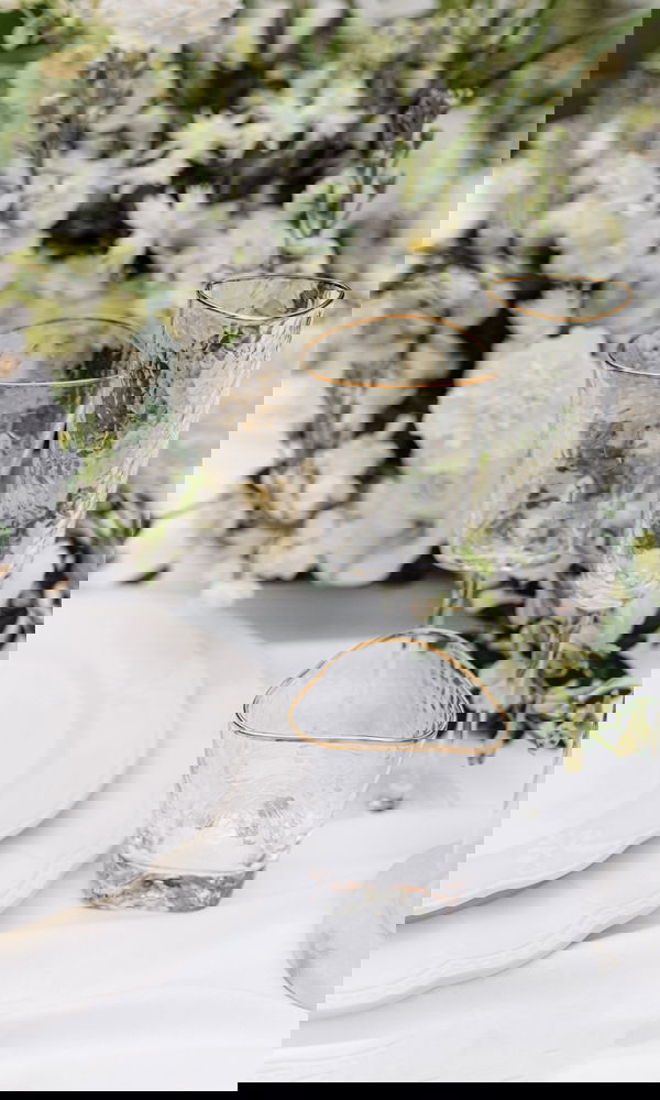 Fein gearbeitete Gläser mit Goldrand und weißem Blumenschmuck bilden eine edle Tischdekoration auf einem in Weiß gedeckten Hochzeitstisch.