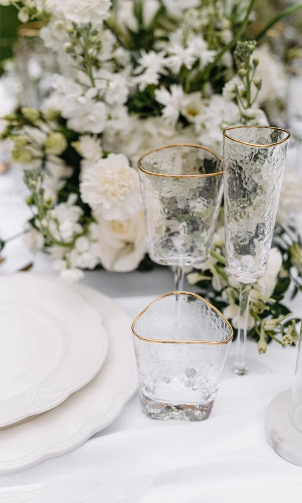 Fein gearbeitete Gläser mit Goldrand und weißem Blumenschmuck bilden eine edle Tischdekoration auf einem in Weiß gedeckten Hochzeitstisch.