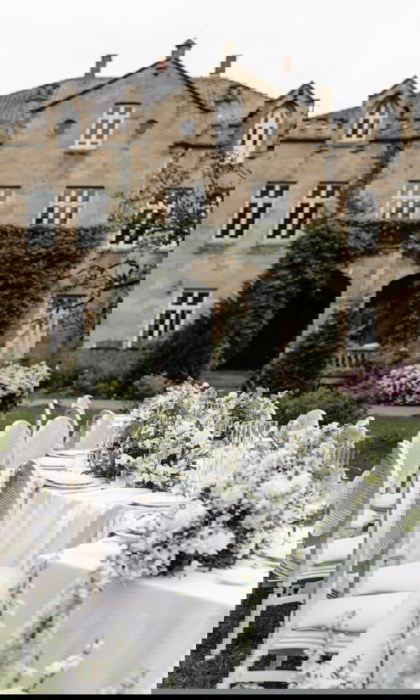 Große, weiß gedeckte Tafel mit Blumenarrangements und klassischen Stühlen im Garten einer historischen Location, bereit für die Hochzeit.
