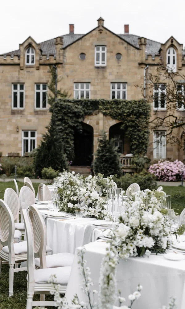 Große, weiß gedeckte Tafel mit Blumenarrangements und klassischen Stühlen im Garten einer historischen Location, bereit für die Hochzeit.