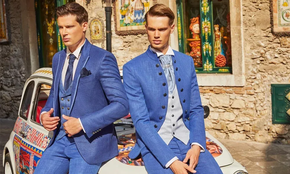 Zwei Männer in eleganten, kräftig blauen Anzügen sitzen auf einer bunt verzierten Autostoßstange vor einer historischen Steinwand; einer trägt einen klassischen Dreiteiler, der andere einen Anzug im festlichen Stehkragenstil mit hellgrauer Weste