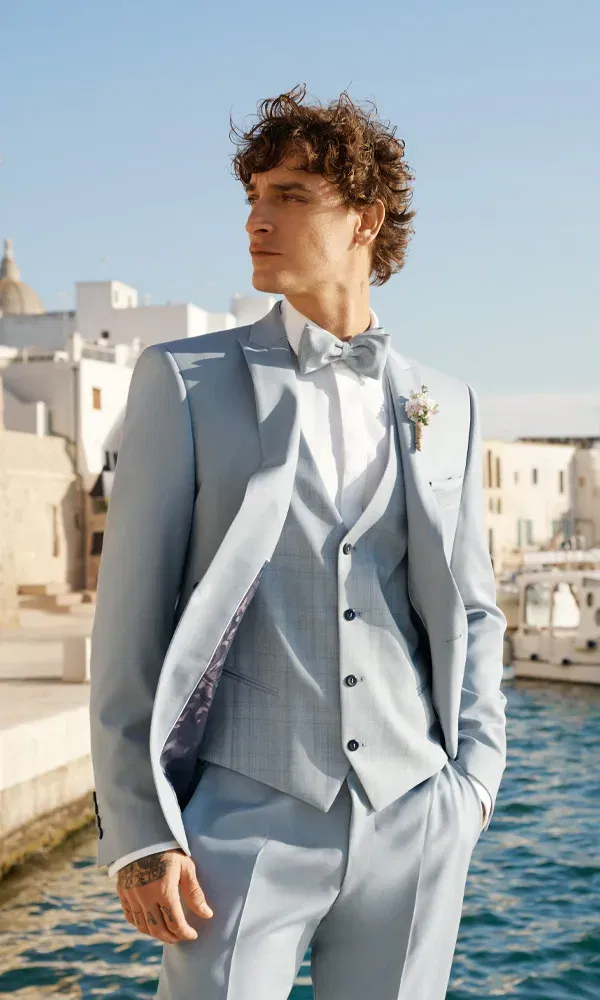 Bräutigam in hellblauem Anzug mit Weste und Fliege steht lässig am Hafen, schaut zur Seite und trägt einen kleinen Anstecker am Revers.