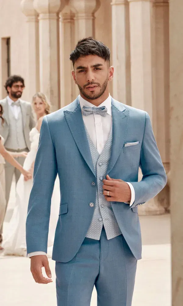Bräutigam in hellblauem Anzug mit gemusterter Weste und Fliege steht vor sandfarbenen Säulen, während im Hintergrund Hochzeitsgäste und ein Brautpaar zu sehen sind.