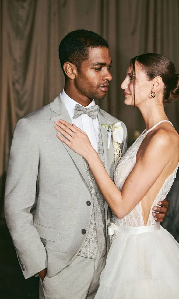 Intimer Moment zwischen Braut und Bräutigam: Er trägt einen hellgrauen Hochzeitsanzug mit Fliege, sie ein elegantes Brautkleid mit tiefem Rückenausschnitt.