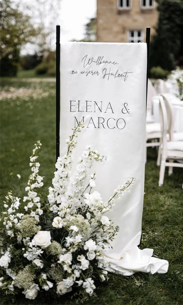 Ein Willkommensschild mit den Namen „Elena & Marco“ sowie weißen Blumenarrangements lädt die Gäste zur Hochzeitsfeier im Freien ein.