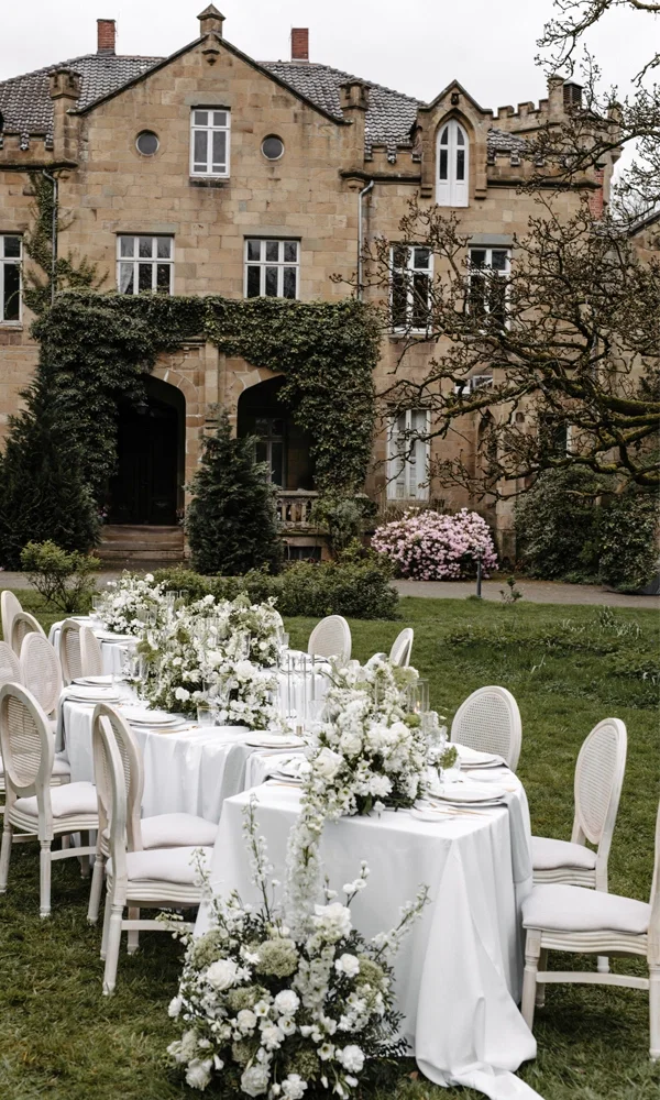Romantische Outdoor-Tafel mit weißen Blumen und gedeckten Stühlen, stilvoll arrangiert im Garten einer historischen Villa.