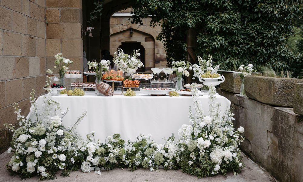 Elegantes Hochzeitsbuffet, dekoriert mit weißen Blumen und frischen Früchten, bereitgestellt auf einem in Weiß gedeckten Tisch in rustikaler Umgebung.