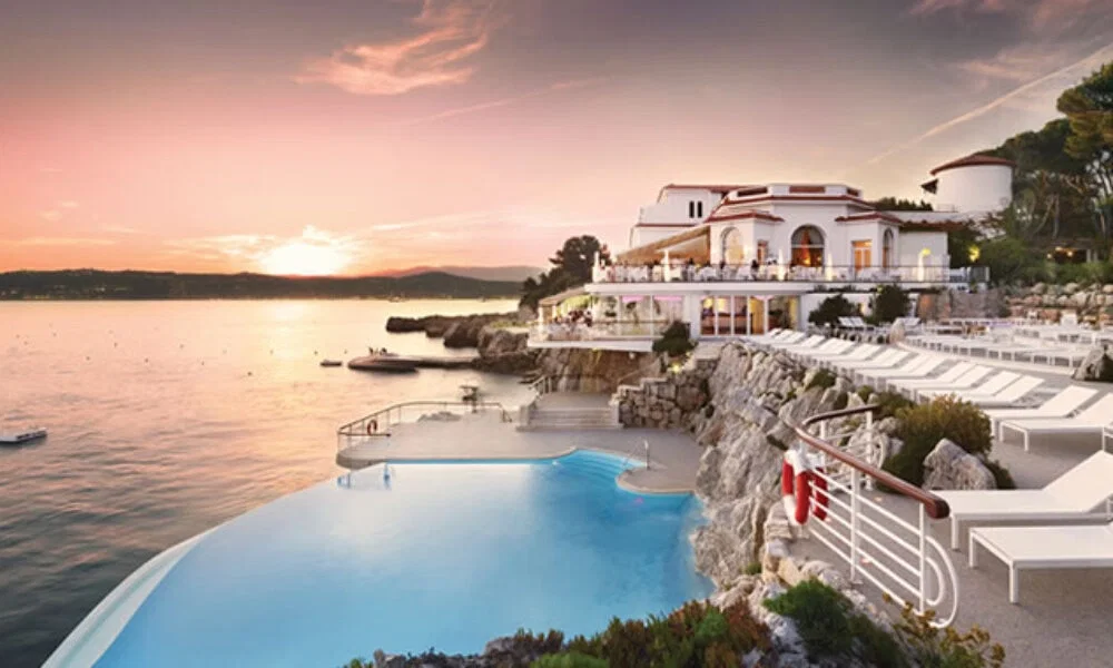 Eleganter Pool in Südfrankreich, eingebettet in Felsen und mit Blick auf das Meer, umgeben von Liegen und einem mediterranen Hotel bei Dämmerung.