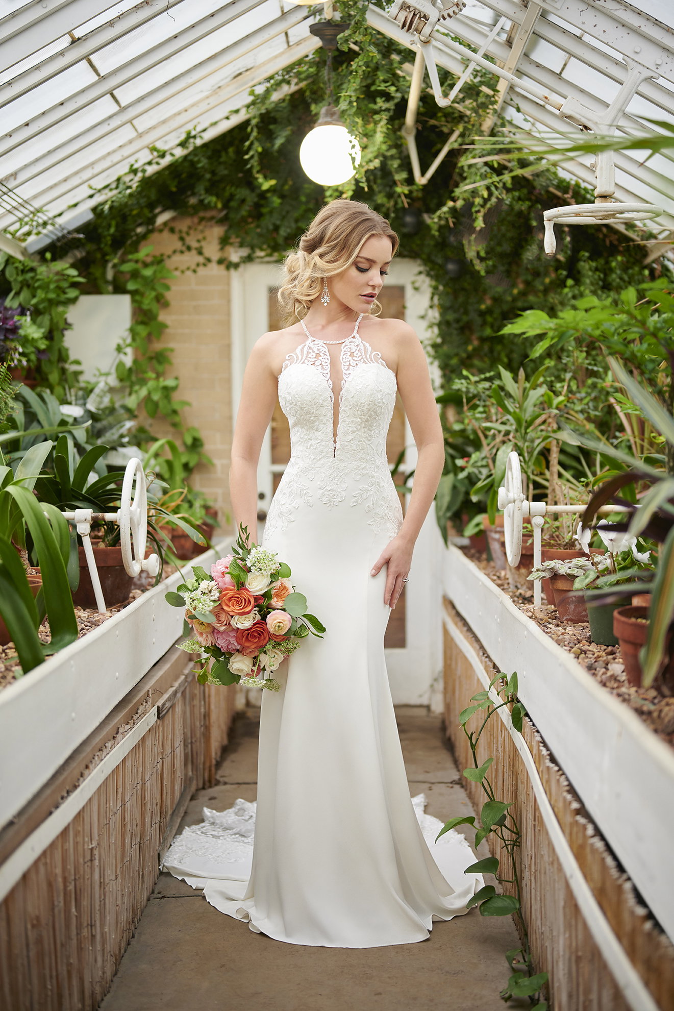 Romantisches Brautbild: Die Braut im bestickten Brautkleid mit transparentem Dekolleté steht in einem Gewächshaus, die Pflanzen im Hintergrund verstärken die natürliche Atmosphäre. Sie trägt einen bunten Blumenstrauß in warmen Tönen.