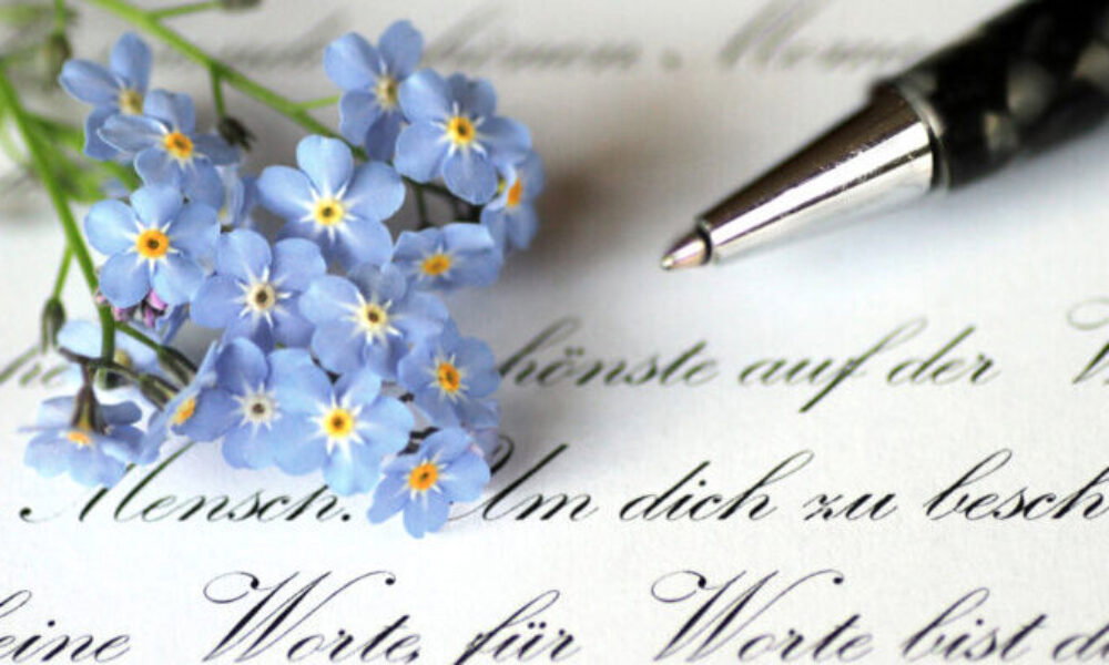 Kleine, blaue Vergissmeinnicht-Blumen liegen neben einem silber-schwarzen Kugelschreiber auf einem Papier, das mit geschwungenen Buchstaben von Hand beschrieben ist.