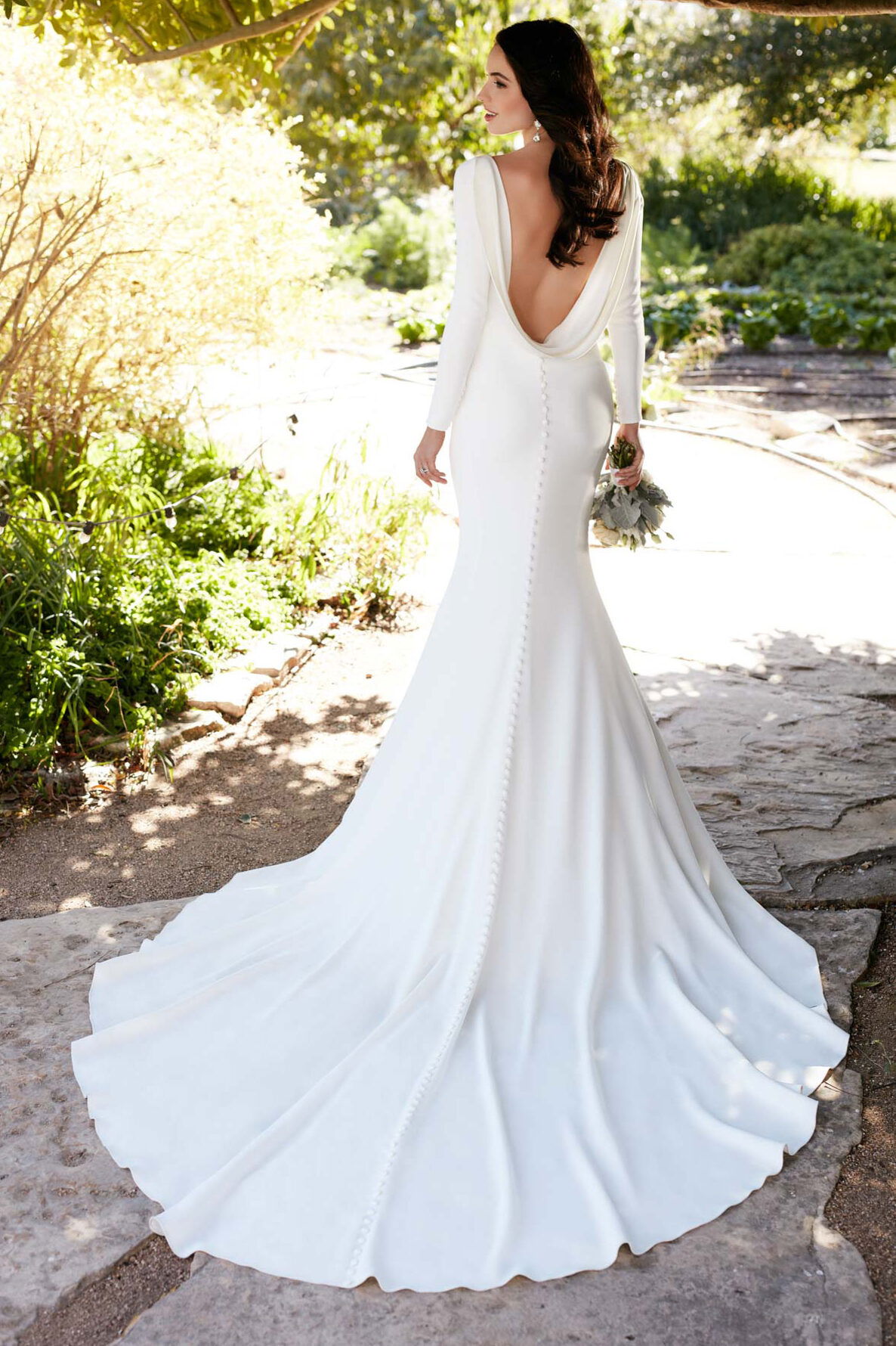 Elegante Braut in einem weißen Kleid mit offenen Rücken und langer Schleppe. Sie steht auf einem Naturweg, umgeben von grünem Laub und Sonnenstrahlen.