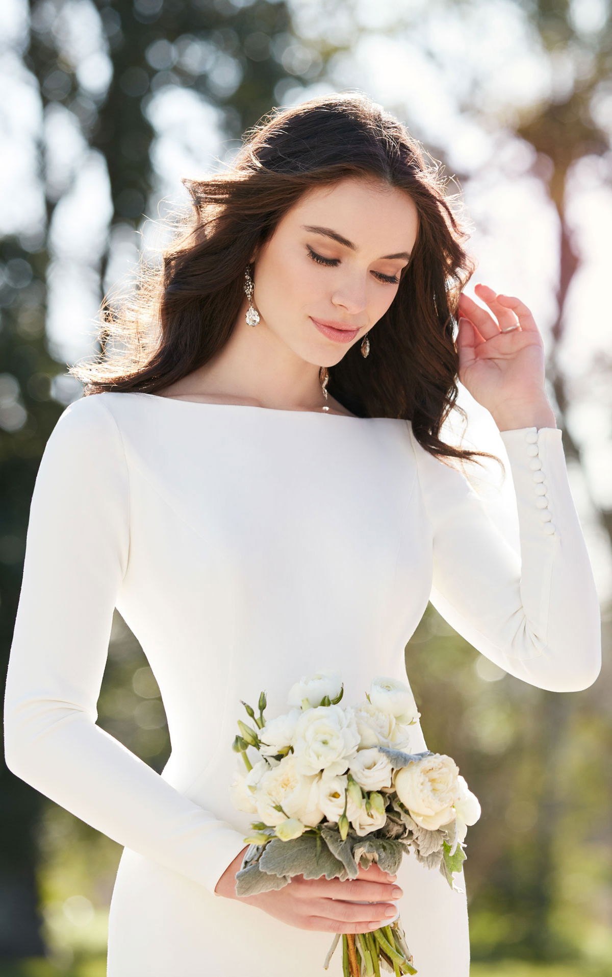 Die Braut trägt ein schlichtes Hochzeitskleid mit langen Ärmeln und einem Bouquet aus weißen Blumen. Sie blickt sanft nach unten, während das Sonnenlicht ihr Haar und Gesicht zart umspielt.