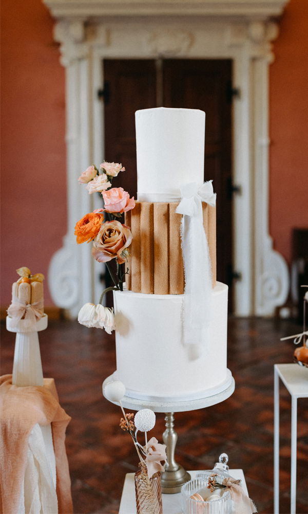 Ein stilvoller Hochzeitstorten-Entwurf, der aus schlichten, weißen Ebenen und Akzenten in Form von getrockneten Rosen besteht. Die Torte steht in einem edlen Raum mit detaillierten Verzierungen im Hintergrund.