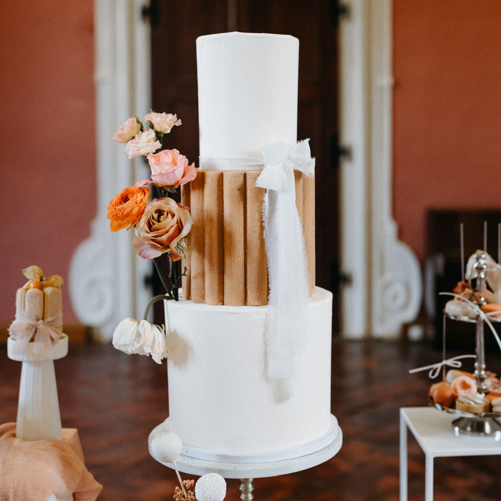 Schlichter Hochzeitskuchen mit einer Kombination aus weißen Ebenen und einem dekorativen braunen Akzent, umgeben von rosafarbenen und orangefarbenen Blumen. Die Torte ist in einem klassischen Saal mit Holzdetails und sanftem Licht platziert.