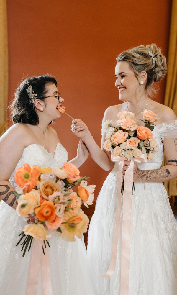 Zwei Frauen in weißen Brautkleidern lachen zusammen und halten üppige Blumensträuße in Orangetönen. Eine Braut bietet der anderen eine Blume an, während sie vor einem warmen, rötlichen Hintergrund posieren.