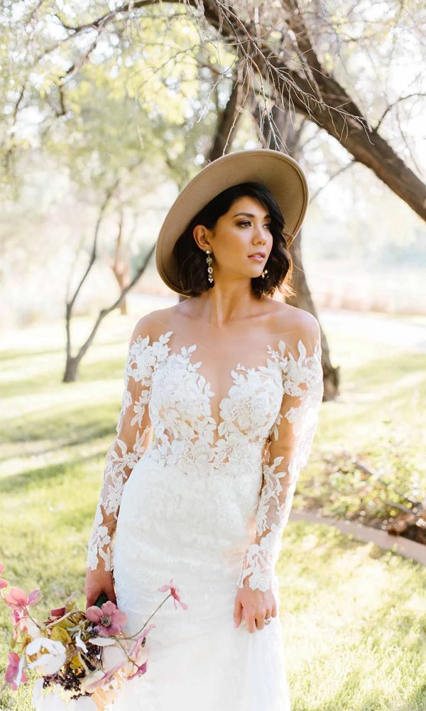 In einem romantischen Spitzenkleid mit floralen Applikationen steht die Braut mit einem Hut und einem Strauß Blumen im Freien. Ihre langen Ärmel und der luftige Schnitt verleihen einen Boho-Look.