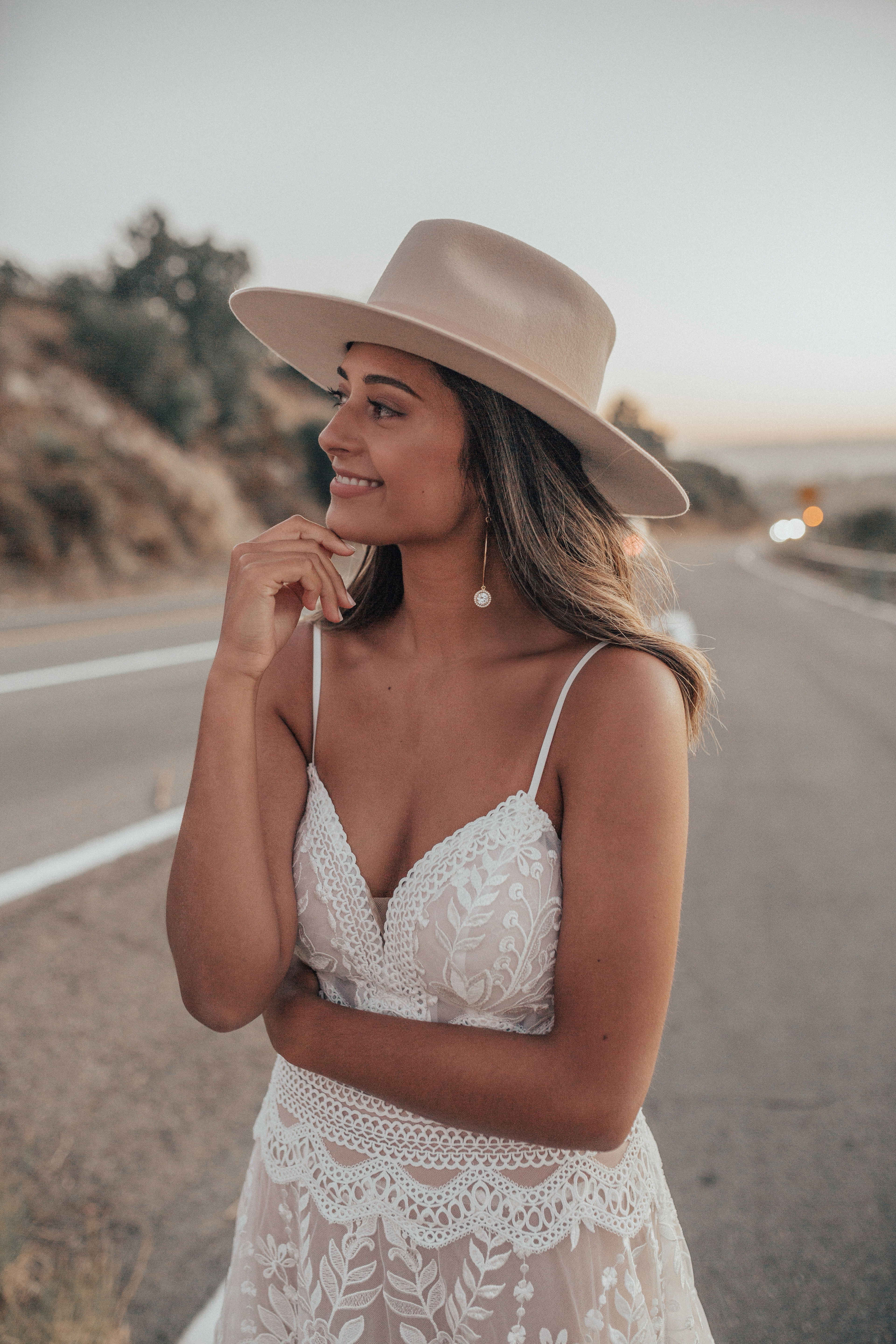 Eine Bräutigam mit Hut in einem boho-inspirierten Spitzenkleid, das die Abendsonne auf dem Highway einfängt und den Horizont betrachtet.