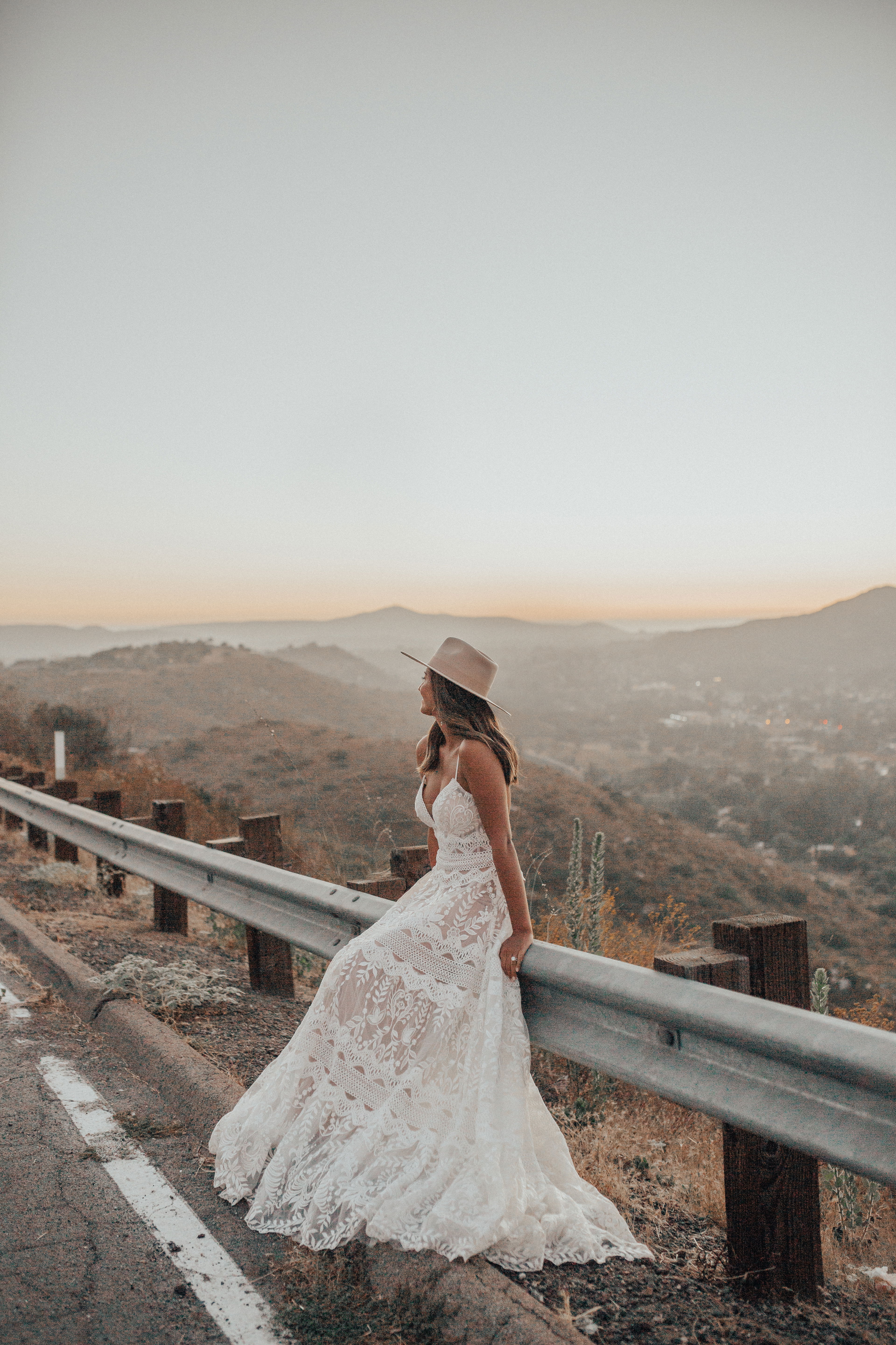 Eine Braut im romantischen Boho-Spitzenkleid sitzt auf einer Leitplanke und lässt den Blick über das Tal bei Sonnenuntergang schweifen.