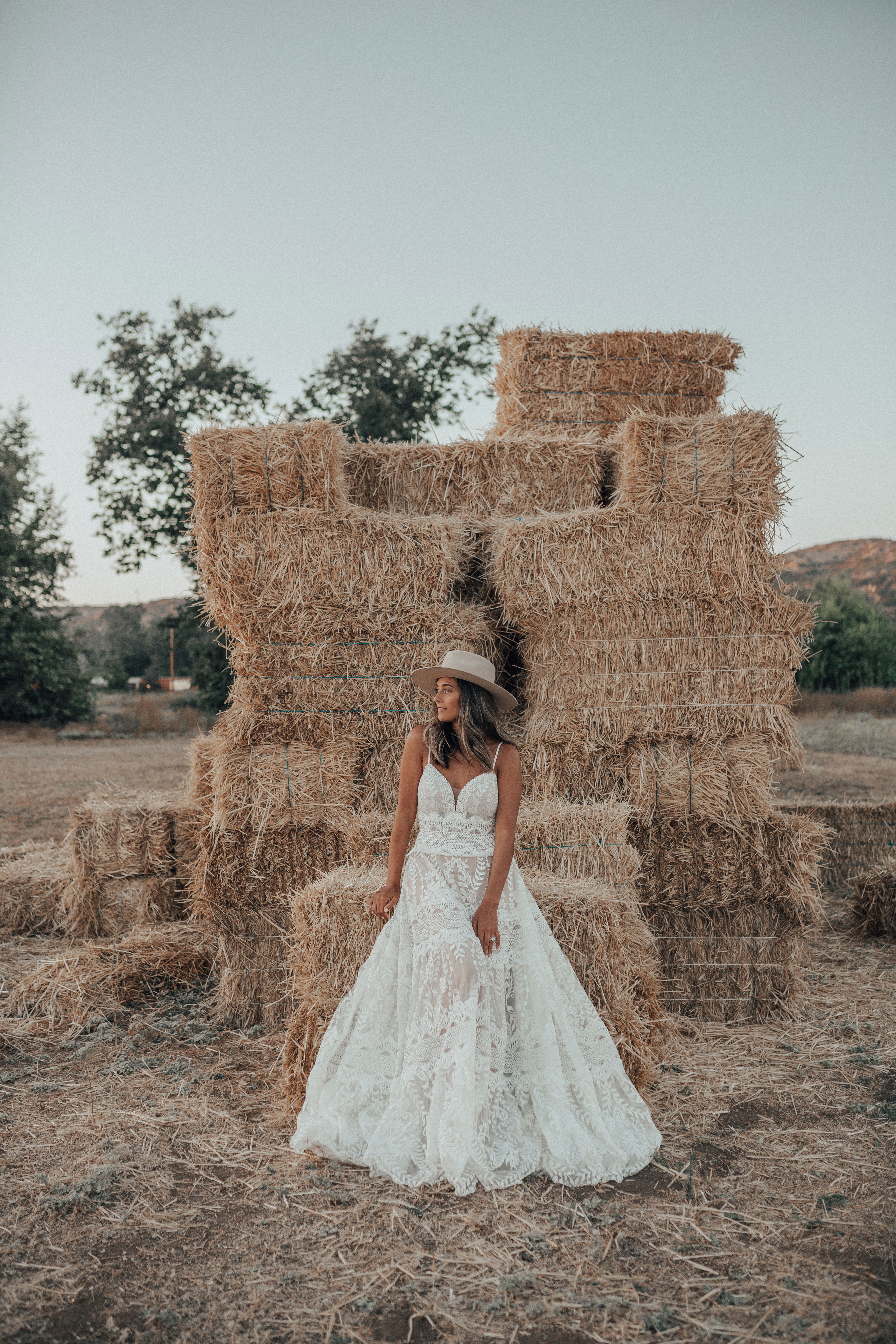 Frau in einem detailreichen, boho-inspirierten Hochzeitskleid posiert auf einem Feld vor Heuballen. Der Strohhut und das ländliche Setting verleihen der Szene eine entspannte Atmosphäre.