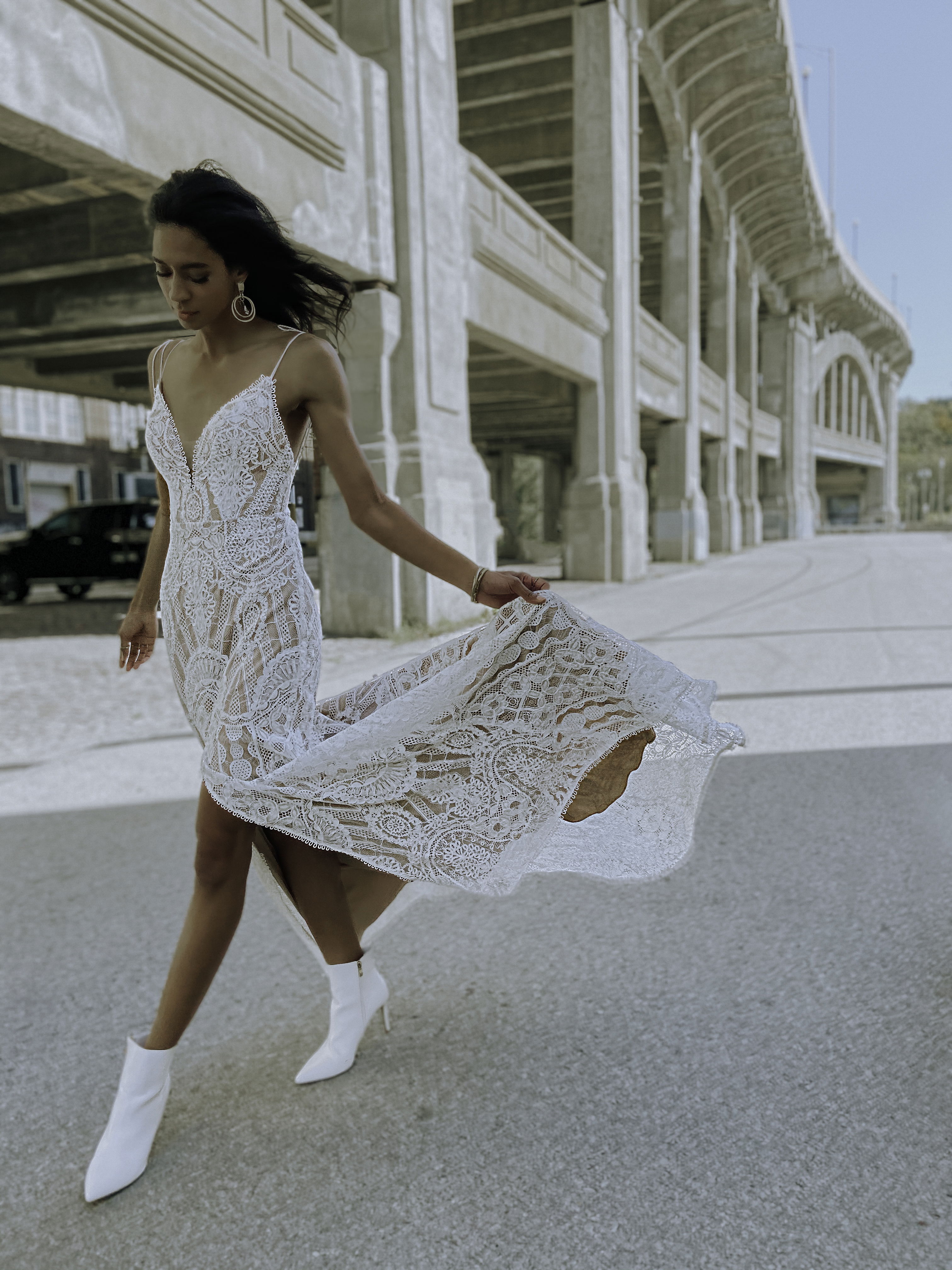 Boho-Braut in einem fließenden Spitzenkleid und weißen Stiefeletten, die dynamisch in einer urbanen Umgebung posiert.