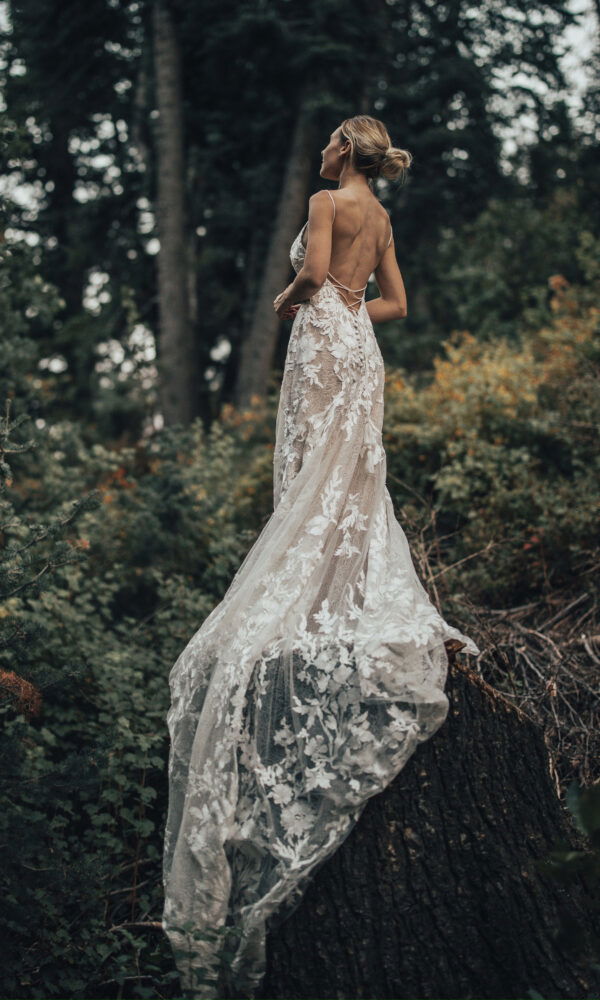 Eine Braut mit einem transparenten Spitzenkleid steht erhöht auf einem Baumstamm, umgeben von üppiger Waldlandschaft, und schaut in die Ferne.