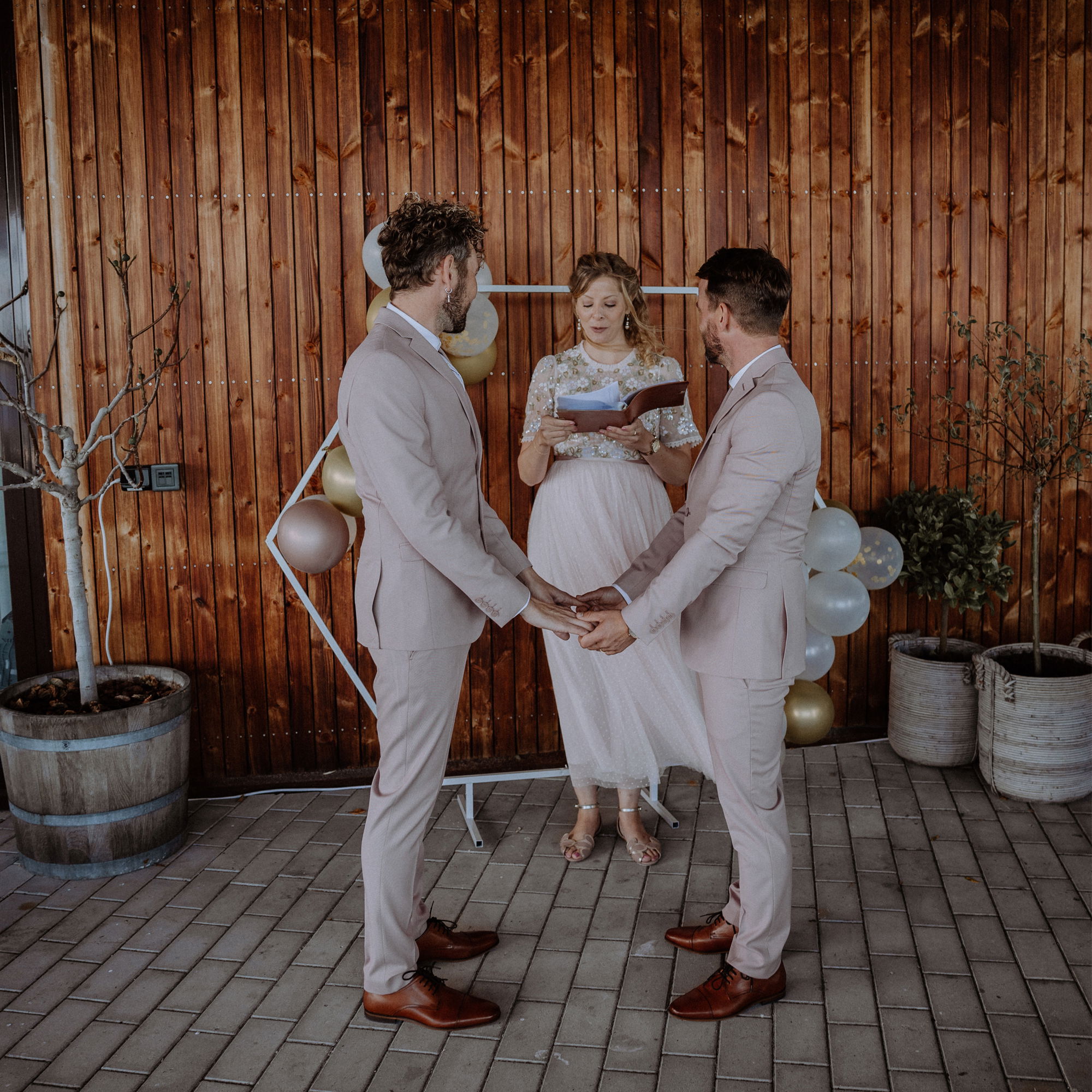 Ein Hochzeitspaar in passenden Anzügen während einer Trauzeremonie, geführt von einer Rednerin mit festlicher Dekoration im Hintergrund.