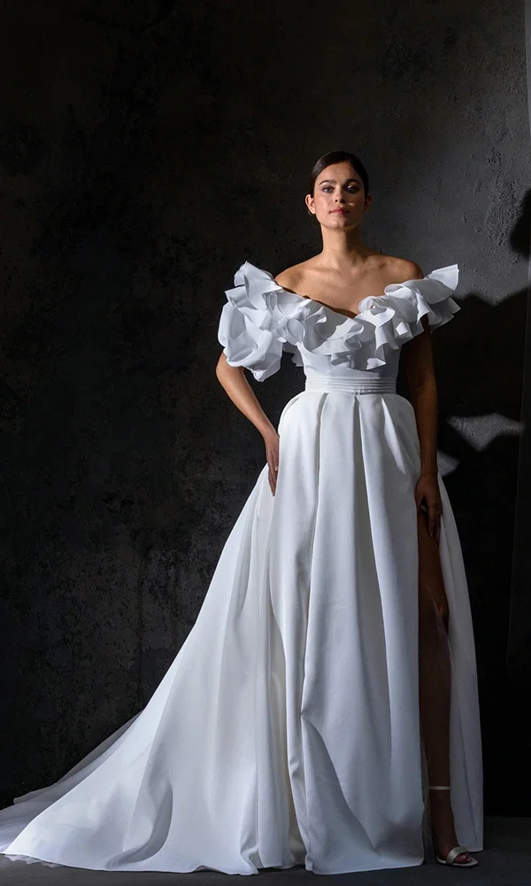 Braut in einem dramatischen, weißen Hochzeitskleid mit voluminösen Rüschen an den Schultern, posiert vor einem dunklen Hintergrund in einem schicken Studio.