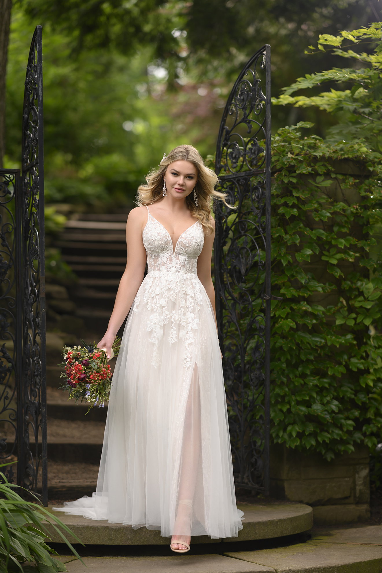 Junge Frau in einem modernen, luftigen Brautkleid mit filigraner Spitze und fließendem Rock, posiert vor einem verzierten Tor inmitten eines malerischen Gartens und hält einen lebendigen Blumenstrauß in der Hand.